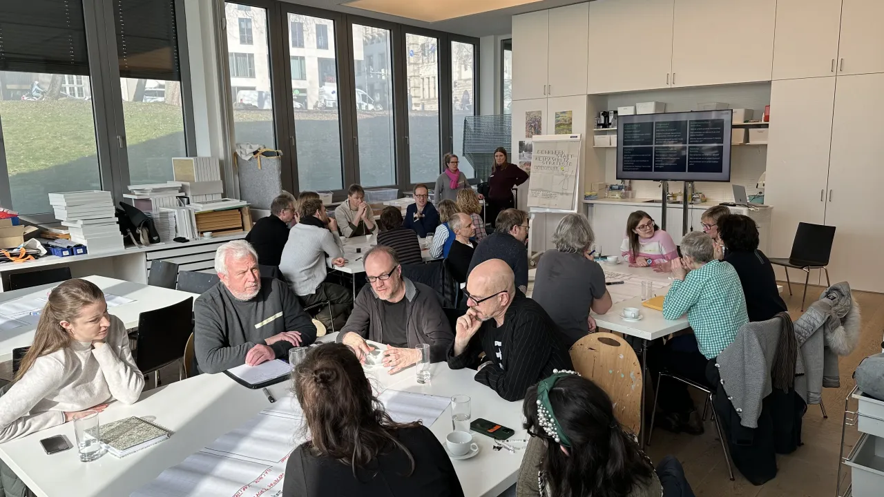 Einige Personen sitzen an Gruppentischen und diskutieren miteinander