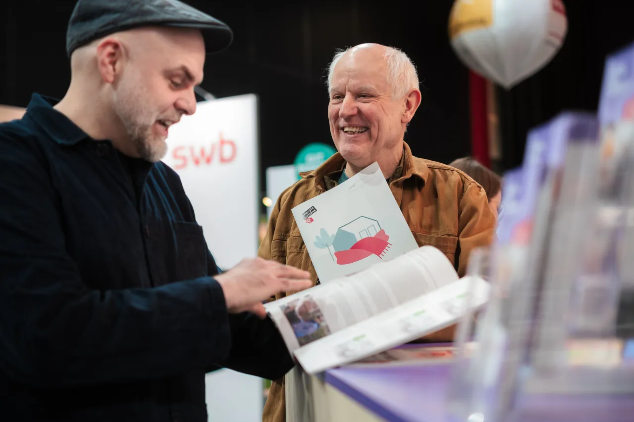 Beratungssituation am Stand des Klima Bau Netzwerks auf den Bremer Altbautagen 2026. Zwei M&auml;nner im Gespr&auml;ch, mit Informationsmaterialien in der Hand.