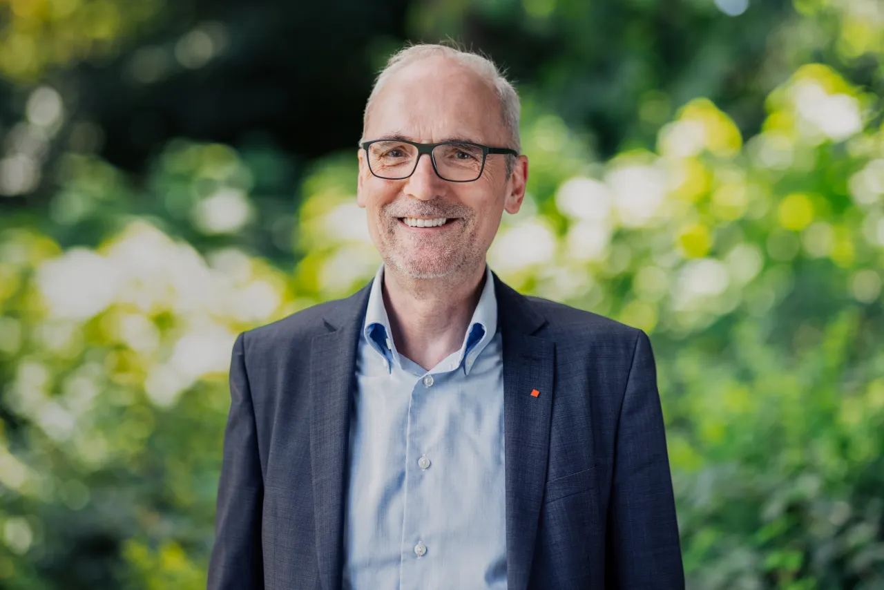 Ein grauhaariger Mann mit Brille lächelt in die Kamera, während er draußen im Grünen steht.