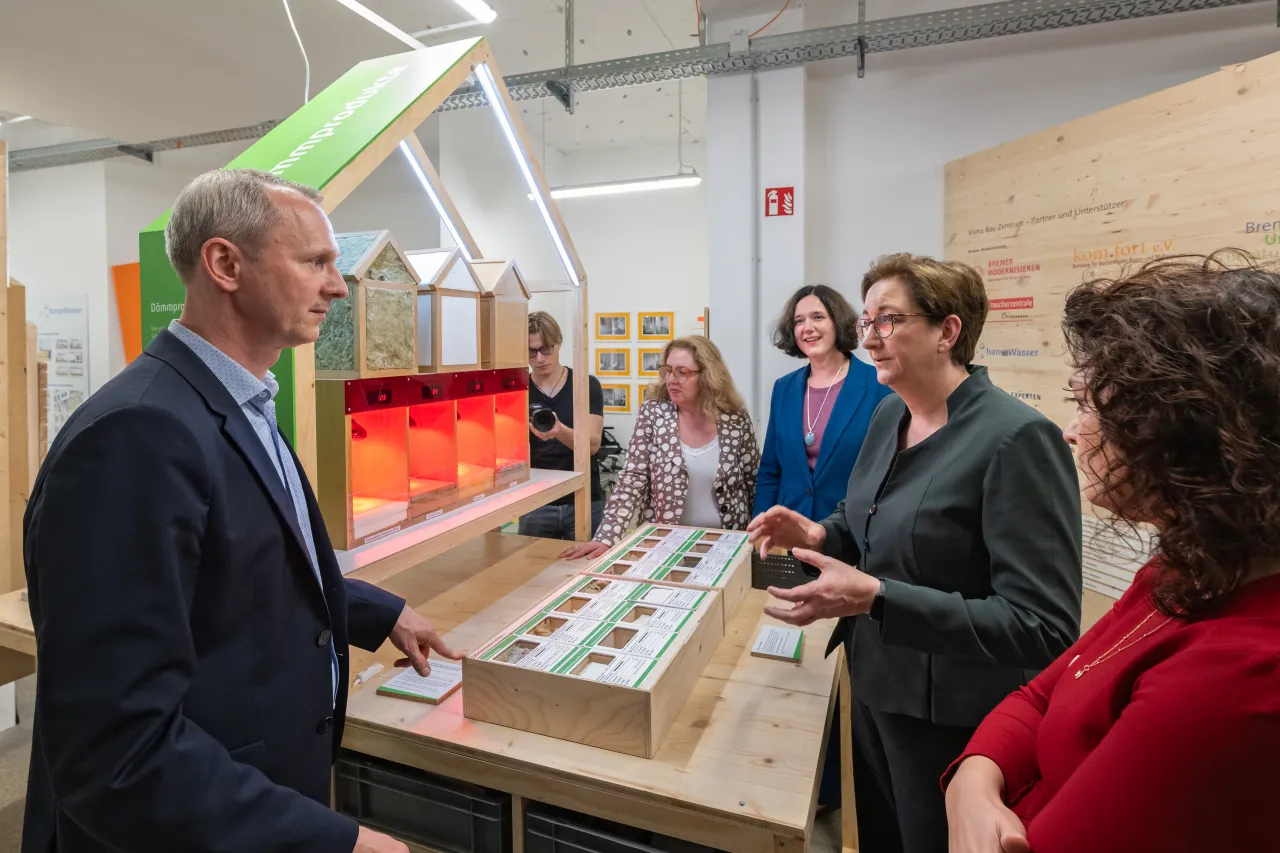 Links Martin Grocholl, rechts stehen Senatorinnen und Bundesbauministerin um Tisch mit Dämmstoffen und leuchtendem Aussttellungsstück.
