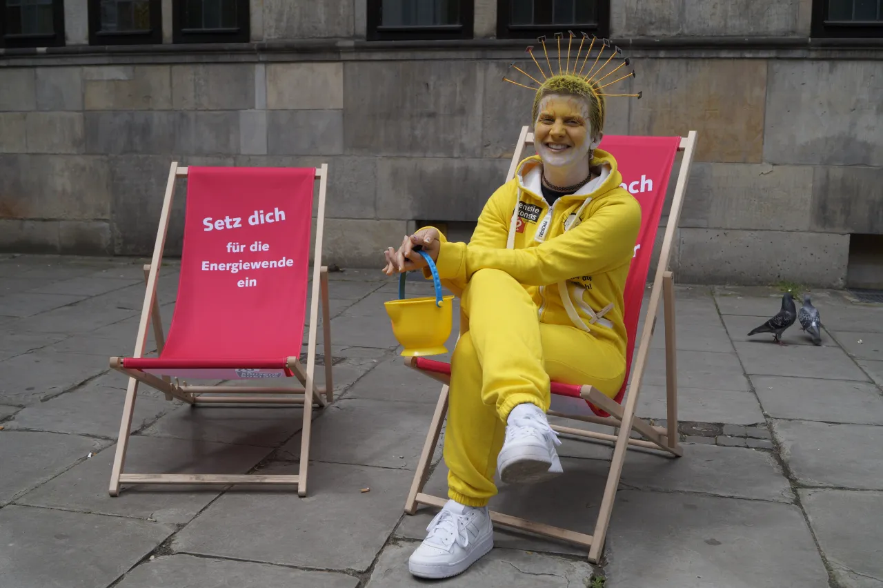 Solarfrau Sunna sitzt in einem Liegestuhl. Deneben steht ein weiterer Liegestuhl mit der Aufschrift 