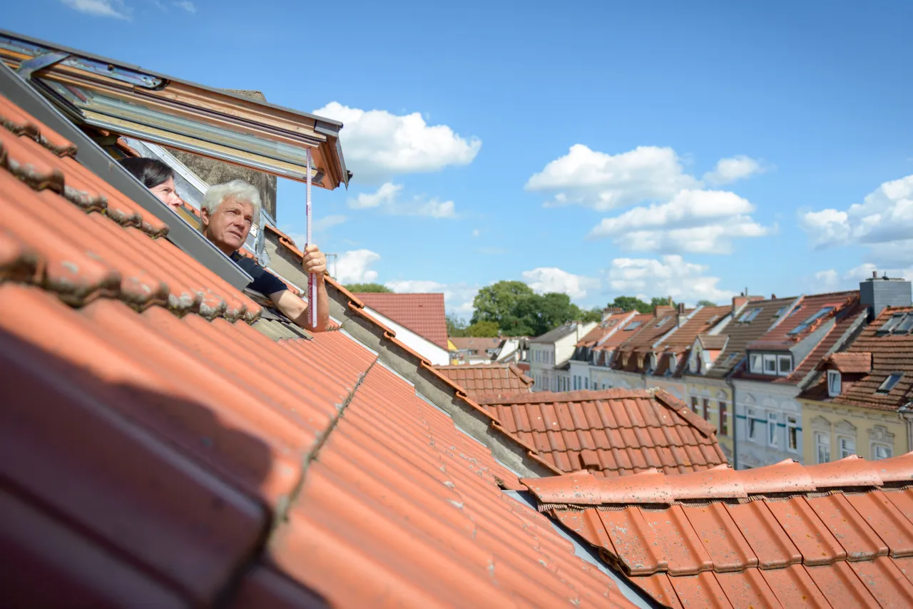Man guckt mit aus Dachfenster und misst mit Zollstock, Gesicht von Frau schaut ebenfalls heraus