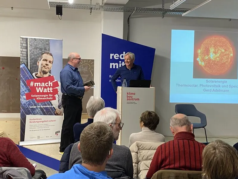 Im Hintergrund stehen zwei Männer auf einer Bühne, im Vordergrund sieht man die Hinterköpfe der Veranstaltungsteilnehmer*innen