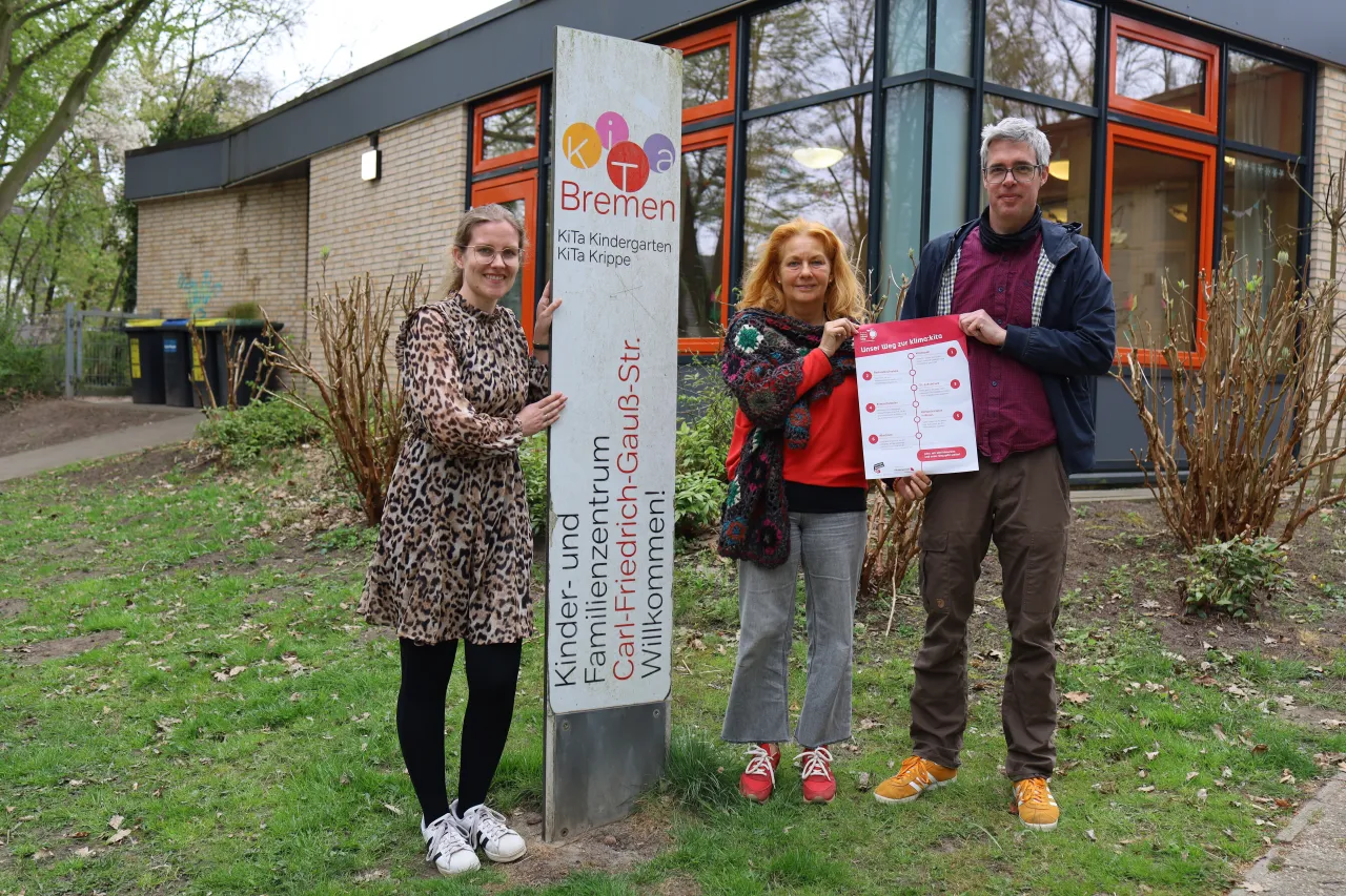 Drei Personen stehen vor einer Kita mit einem Plakat in den Händen.