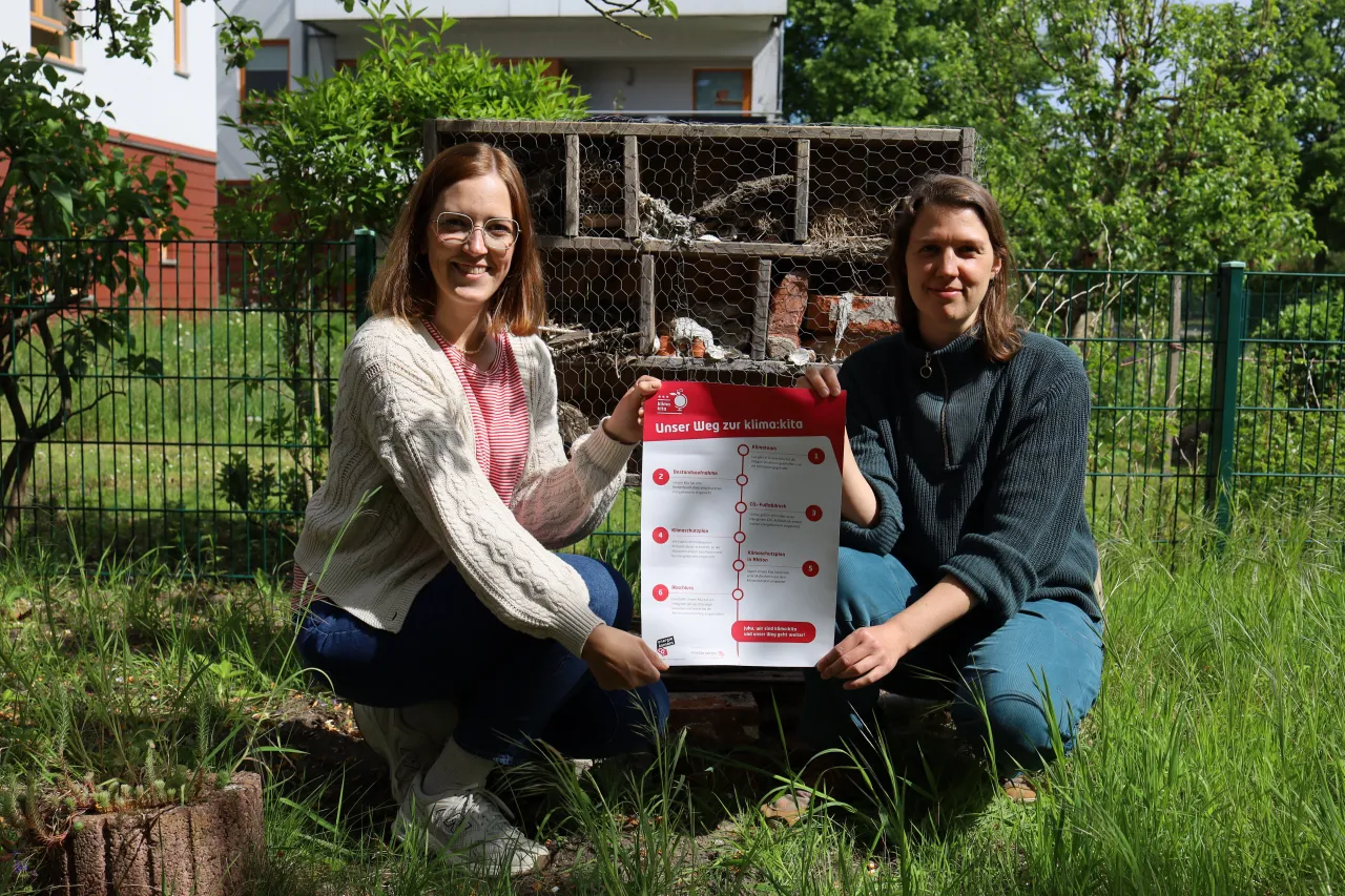 Zwei Frauen mit einem Plakat vor einem Insektenhotel.