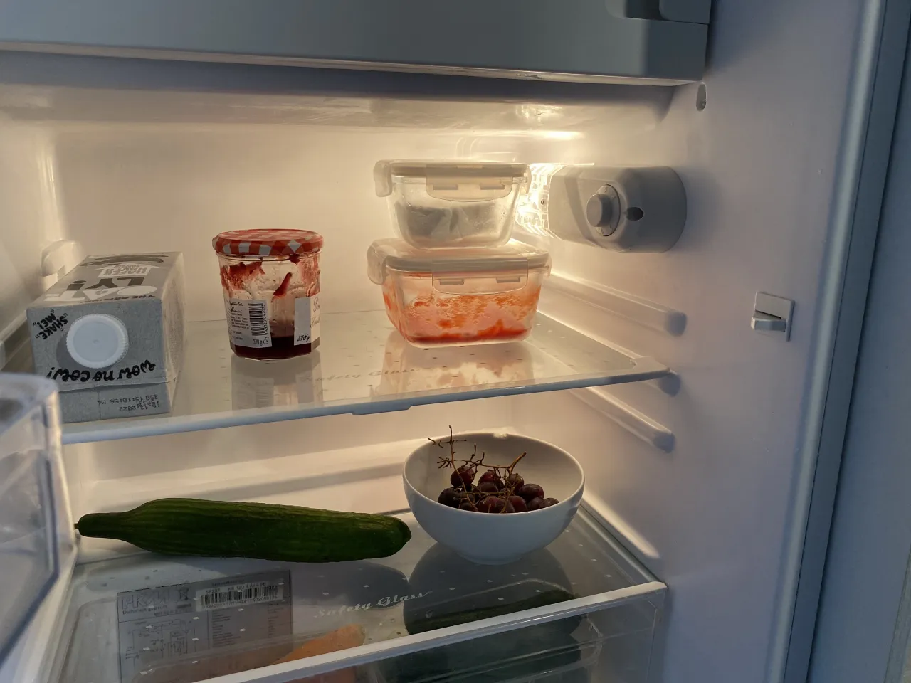 Innenaufnahme von Kühlschrank, gefüllt mit Gurke, Trauben, Hafermiclh, Marmelade und Mehrwegbehältern