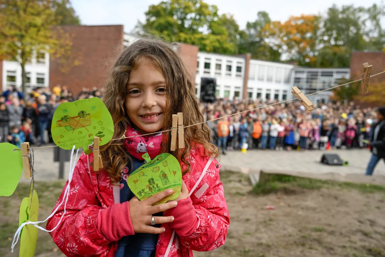 Mädchen hängt einen bemalten Apfel aus Papier an Wäscheleine, Menge an Schülern im Hintergrund
