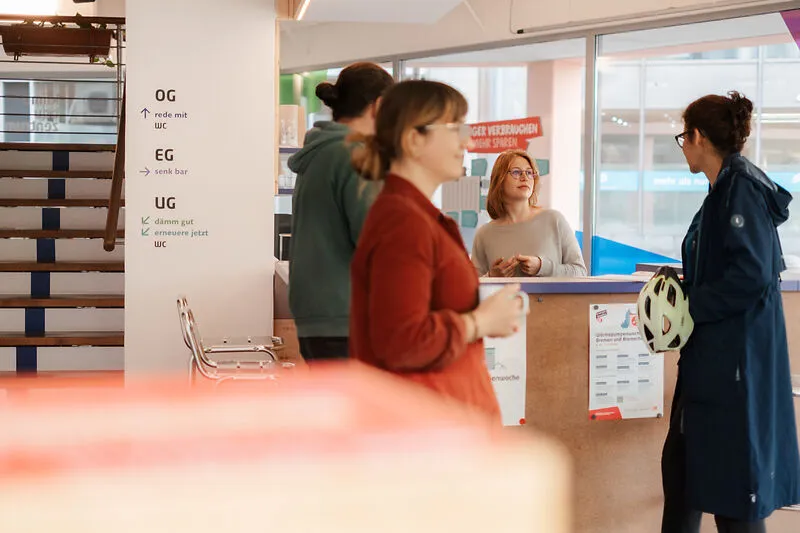 Eingangssituation im Klima Bau Zentrum. Drei Personen stehen am Tresen w&auml;hrend eine vierte Person durchs Bild l&auml;uft.