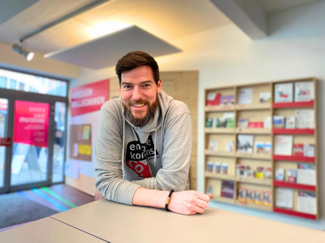 Ein Mann lehnt sich l&auml;chelnd an einen Tresen. Im Hintergrund Infobl&auml;tter in einem Regal.