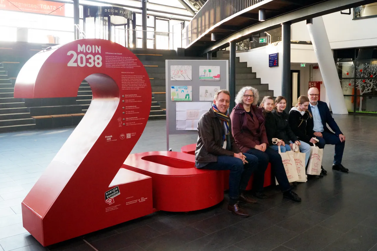 Menschen sitzen auf der Installation Moin 2038 auf der Havenplaza, im Hintergrund Stellwand mit Bildern der Schülerinnen