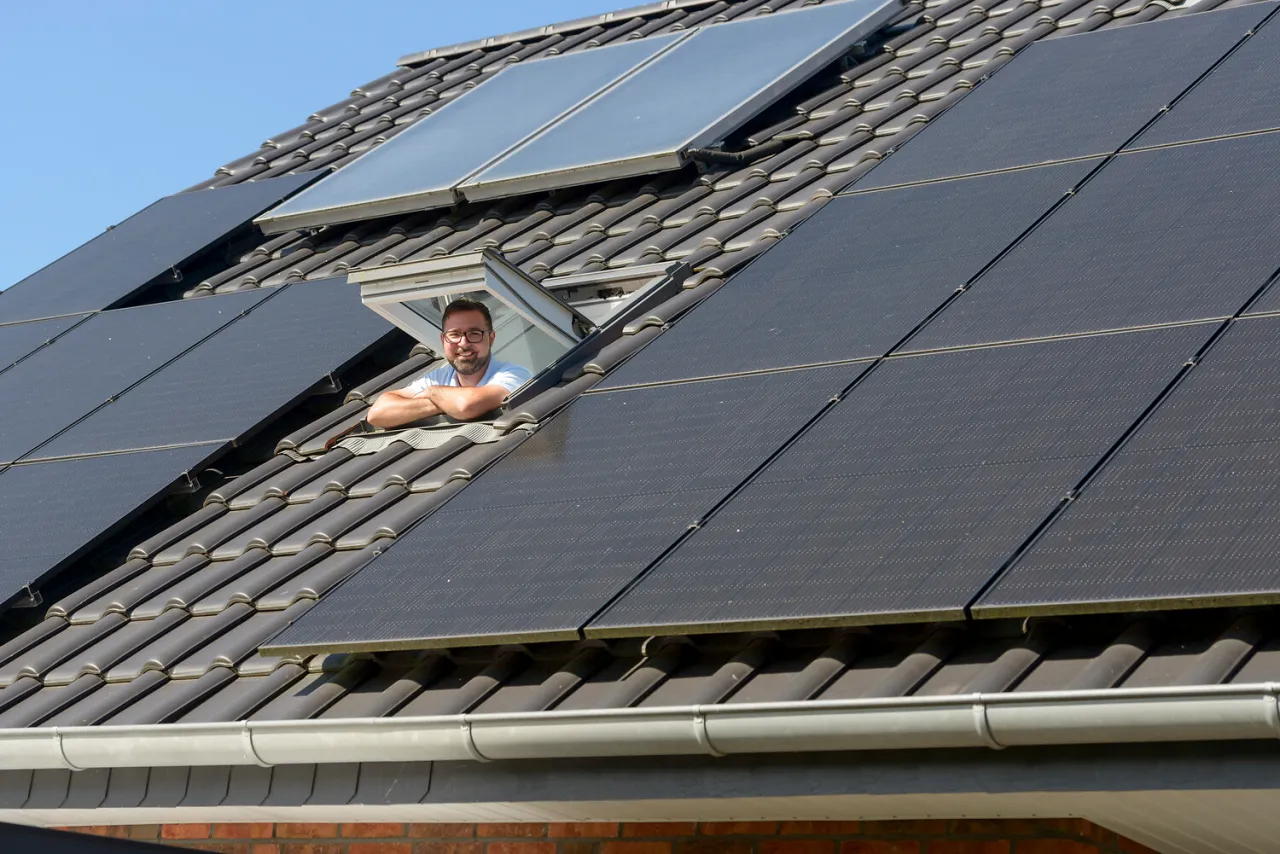 Ein zufriedener Solaranlagen-Besitzer schaut aus einem Dachfenster zwischen Photovoltaikmodulen.