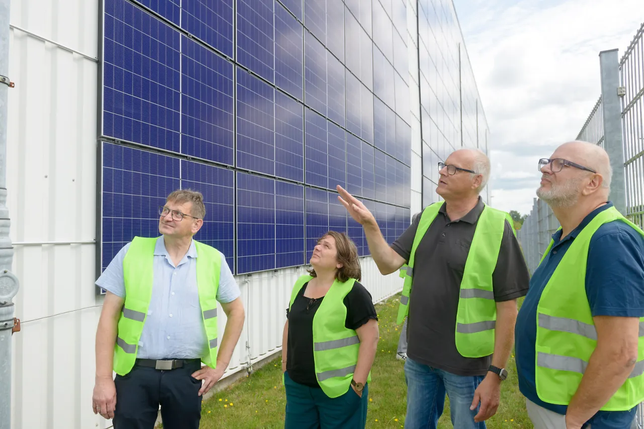 Vier Menschen stehen vor Fabrikhalle, die mit zahlreichen Photovoltaik-Modulen verkleidet ist.