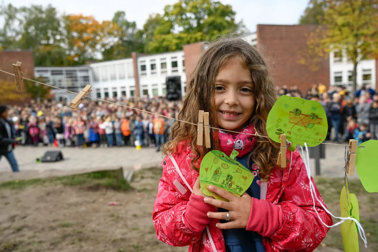 Kind steht vor einer Schule und h&auml;lt einen Apfel aus Papier in der Hand. darauf stehen Klimaschutztipps. Im Hintergrund verschwommen: die Schulgemeinschaft.