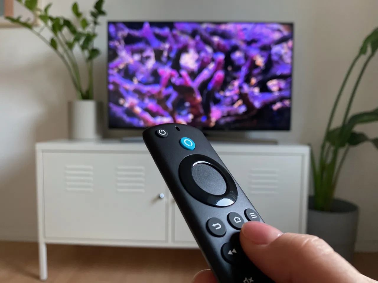 Fernbedienung wir in Hand gehalten, im Hintergrund steht Fernseher mit lilafarbenden Bild