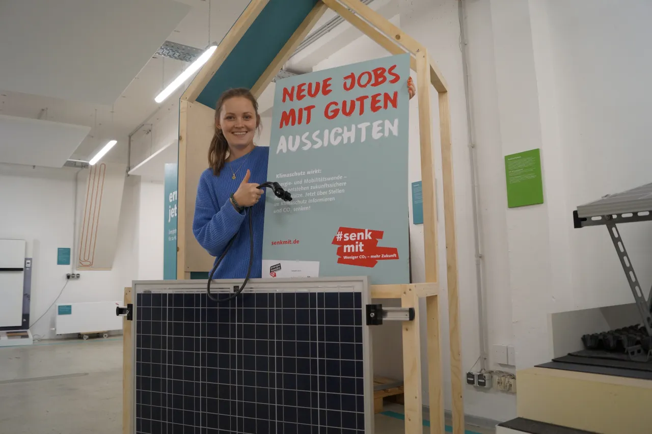 Frau steht hinter Balkonsolarmodul und hält ein Schild, auf dem steht 