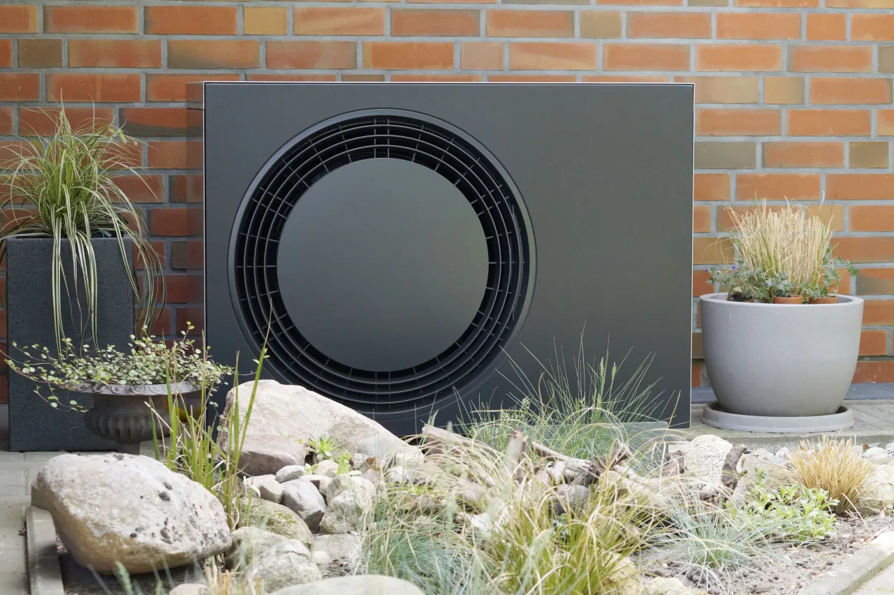 Eine W&auml;rmepumpe steht im Vorgarten vor einer Hauswand, daneben stehen Blument&ouml;pfe.