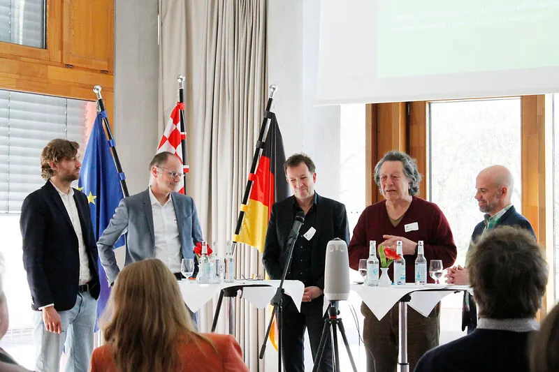 F&uuml;nf Personen diskutieren an Stehtischen vor einem Publikum.