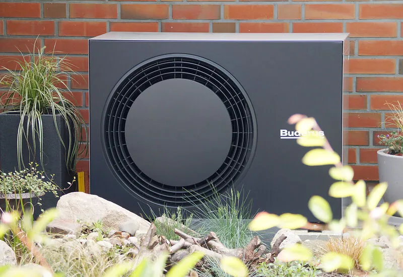 Die dunkelgraue Au&szlig;eneinheit einer W&auml;rmepumpe vor einer Hauswand aus Backstein.