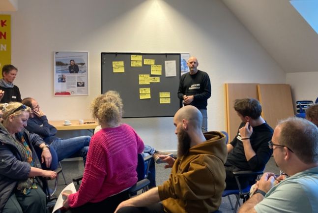 Menschen sitzen in einem Bersprechungsraum und hören einem Mann zu.