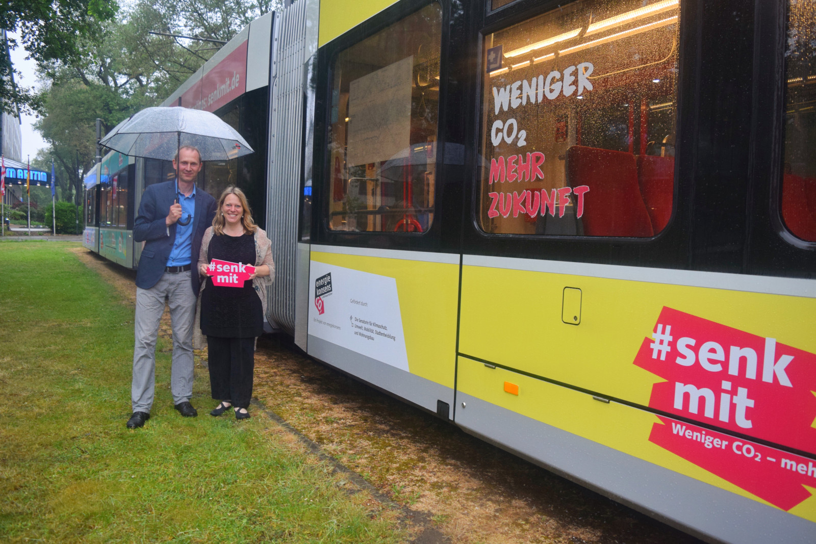 Martin Grocholl und Senatorin Maike Schaefer stehen links neben Straßenbahn im Design der Kampagne #senkmit, Grocholl hält Schirm