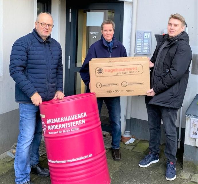 Drei energiekonsens Mitarbeiter*innen stehen vor Tür des neuen Büros und halten Umzugskarton und Tonne mit der Aufschrift