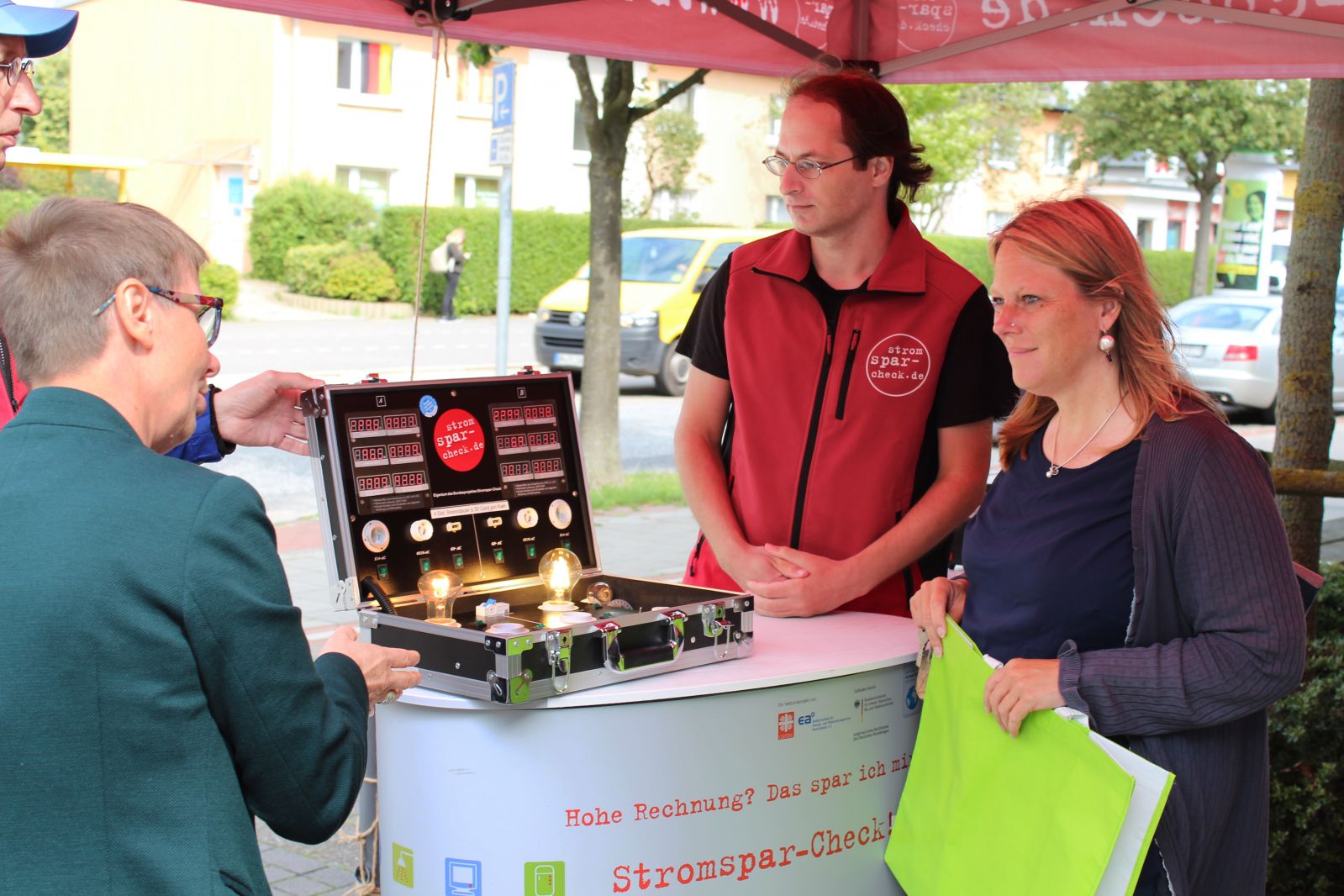 Senatorin Maike Schaefer steht an einem Stand des Stromspar-Checks. Drei Stromsparhelfer*innen demonstrieren mit Hilfe eines Koffers, wie Strom bei Beleuchtung gespart werden kann.