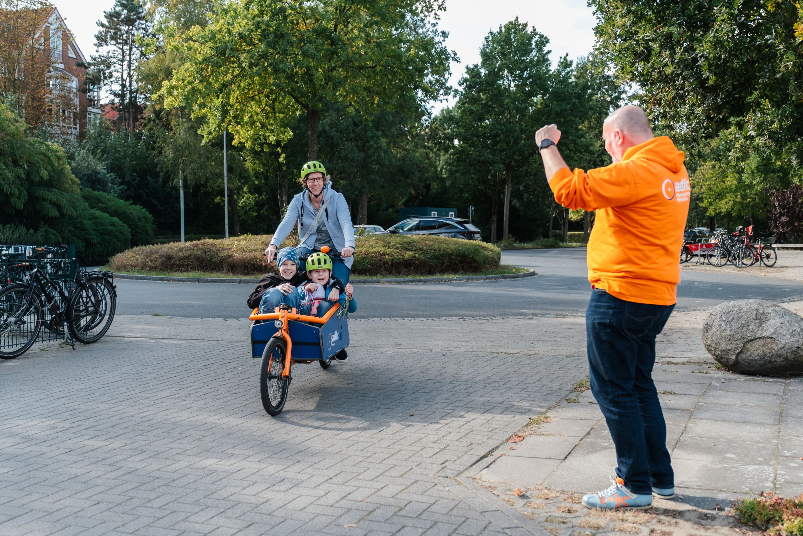 Frau fährt mit zwei Kindern im Lastenrad. Mann vom ADFC freut sich mit ihnen.