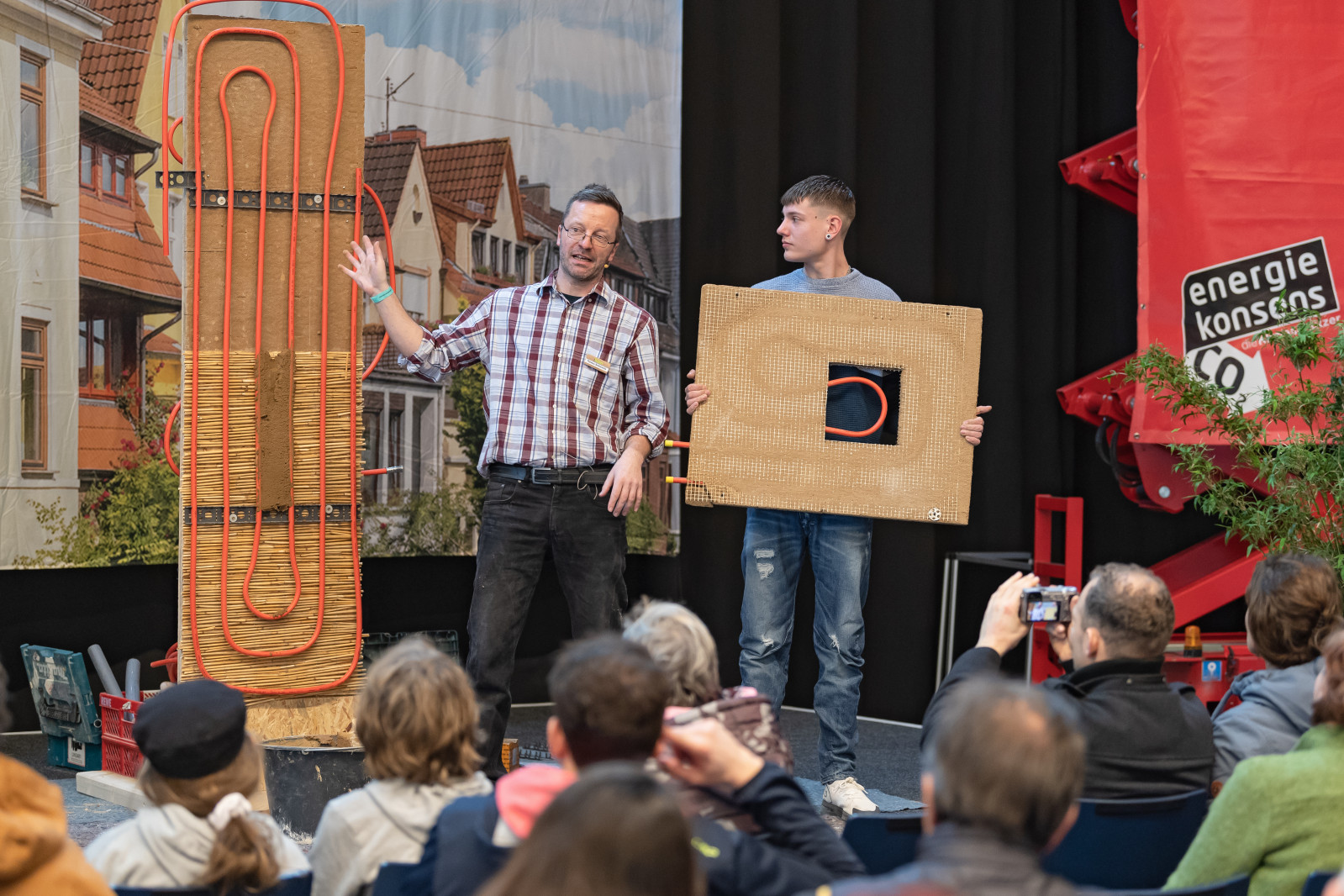 Ein älterer und ein jüngerer Mann stehen mit Ausstellungsstücken von Fußbodenheizung auf der Bühne der Messe Bremer Altbautage und erklären dem Publikum etwas.
