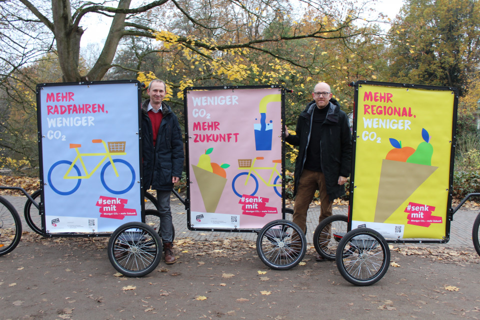 Martin Grocholl und Ronny Meyer stehen zwischen drei Plakat-Rädern der Kampagne #senkmit