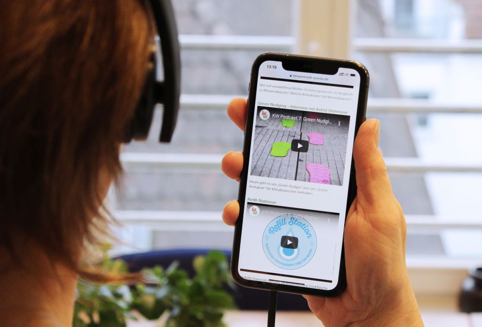 Person mit Kopfhörern hält Smartphone hoch, auf dem ein Podcast läuft