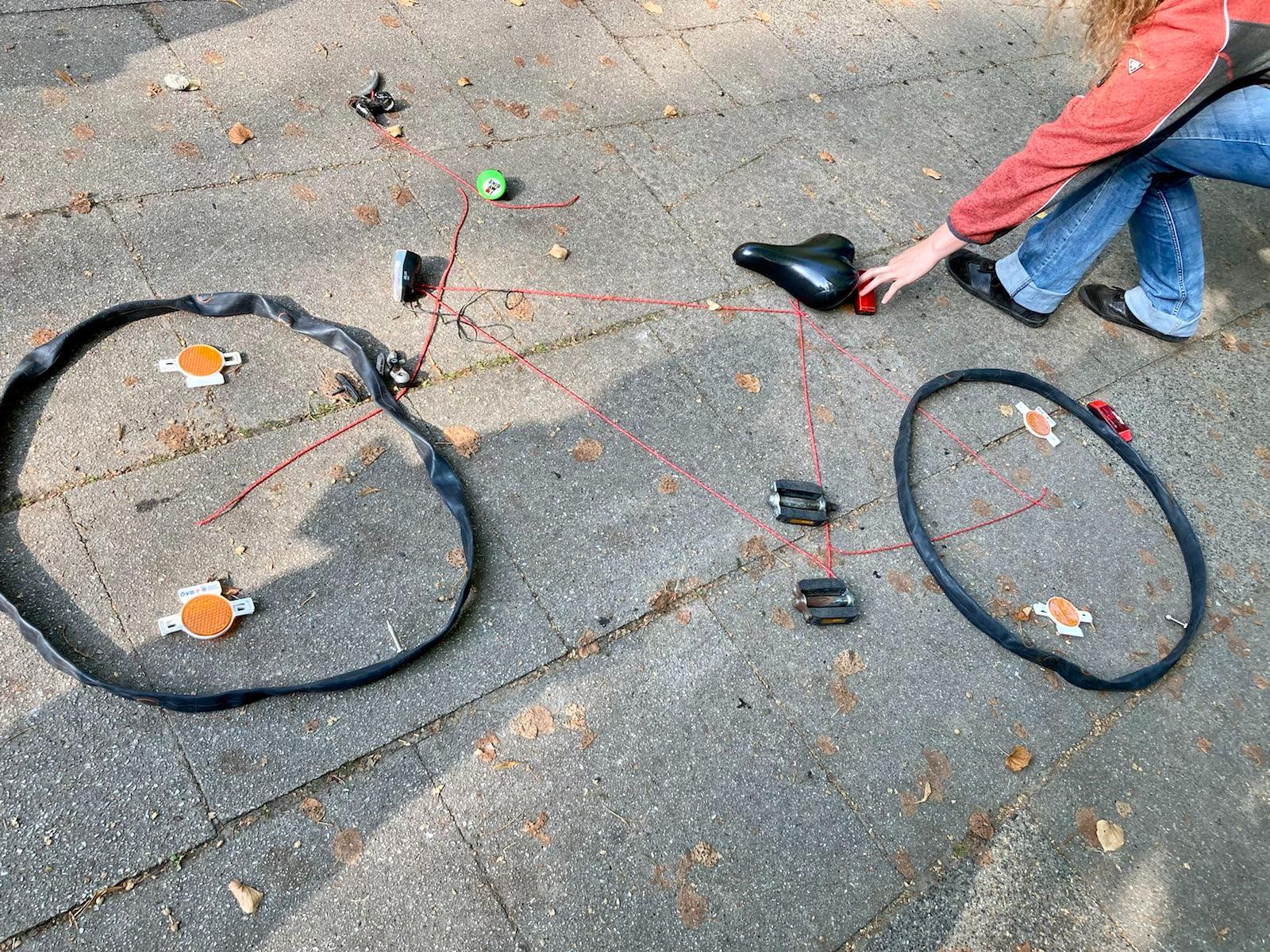 Verschiedene Fahrradteile liegen auf dem Boden und sind mit einem Faden zu einem Fahrrad zusammengelegt
